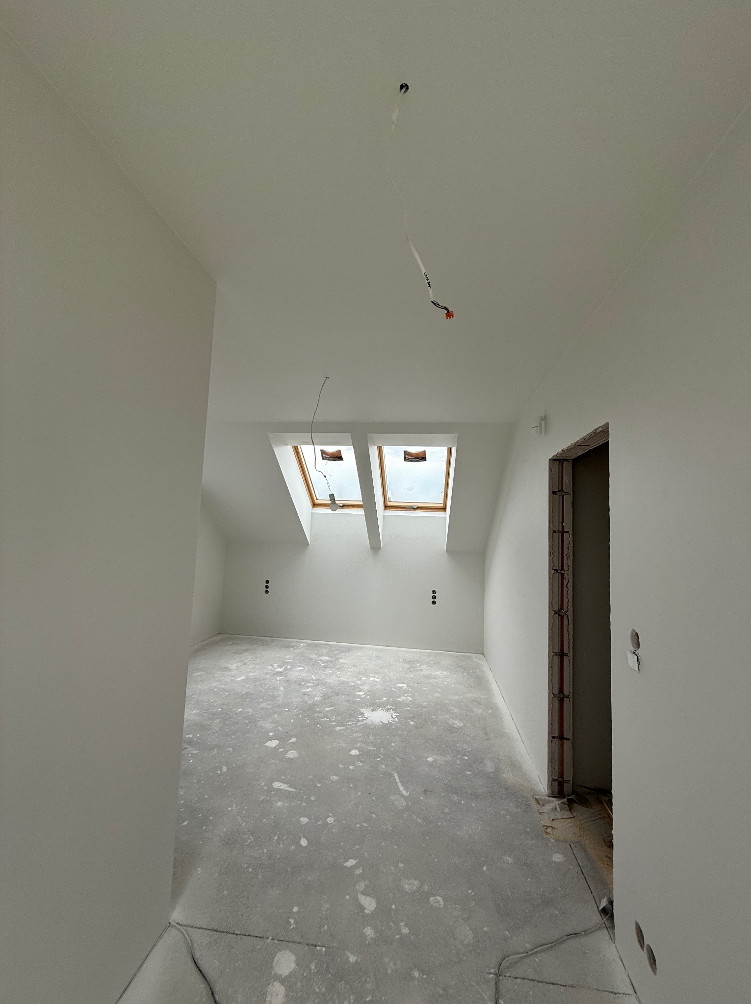 Wnętrze poddasza po remoncie, świeżo pomalowane na biało, z dwoma oknami dachowymi i widocznymi gniazdkami elektrycznymi. Podłoga zabezpieczona folią malarską.