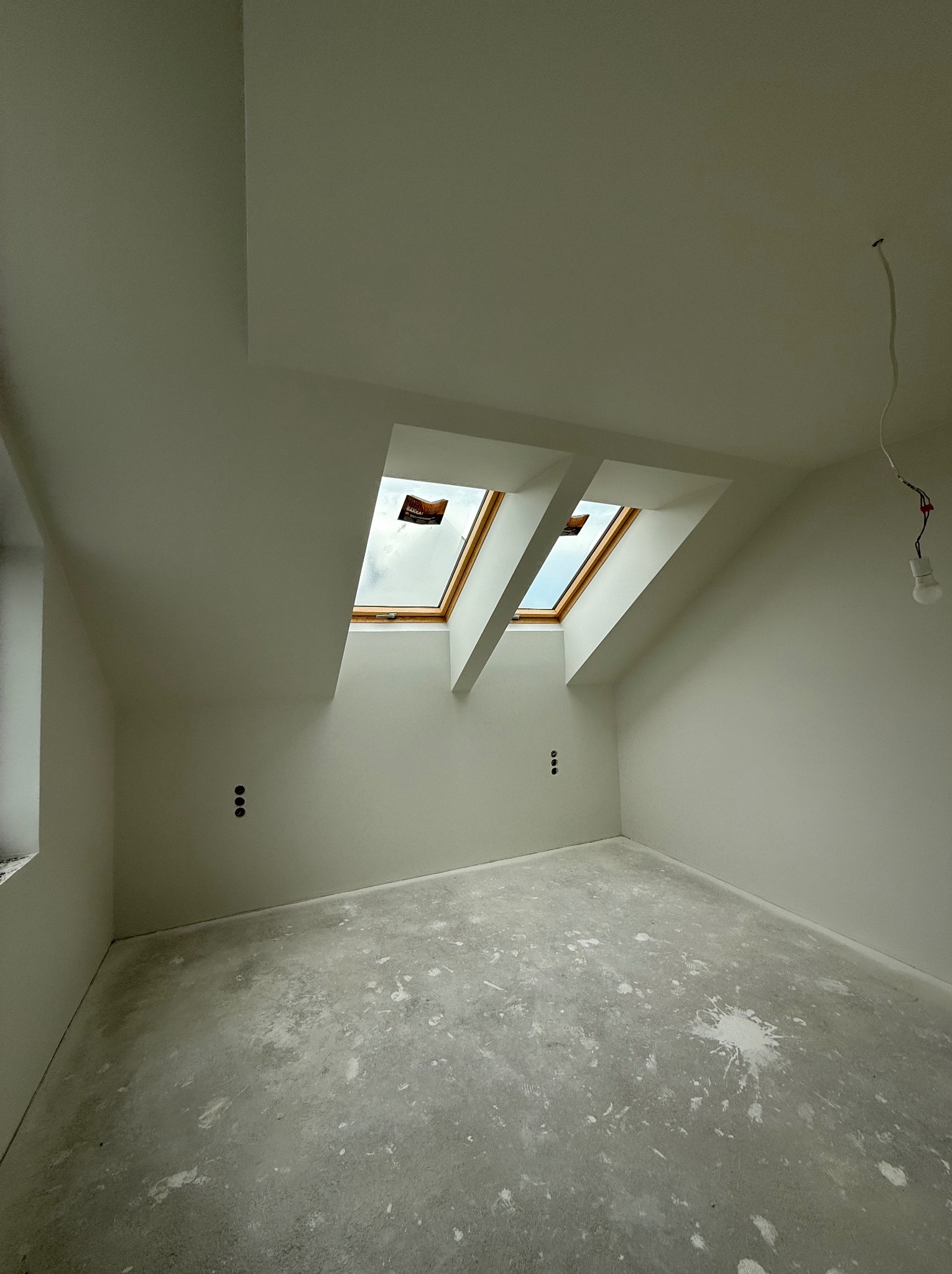 Jasne poddasze z dwoma oknami dachowymi i gładkimi ścianami po szpachlowaniu, przygotowane do malowania. Widoczne puszki elektryczne i surowa podłoga.