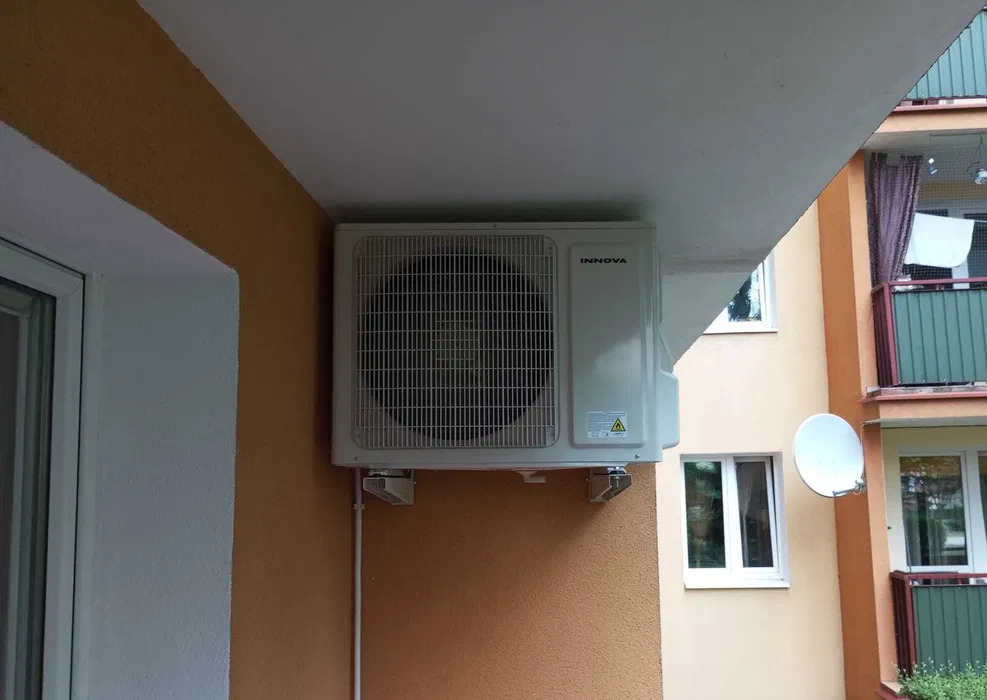 Zewnętrzna jednostka klimatyzatora Innova zamontowana na pomarańczowej ścianie balkonu budynku wielorodzinnego, widoczne wsporniki montażowe i rurka odprowadzająca skropliny.