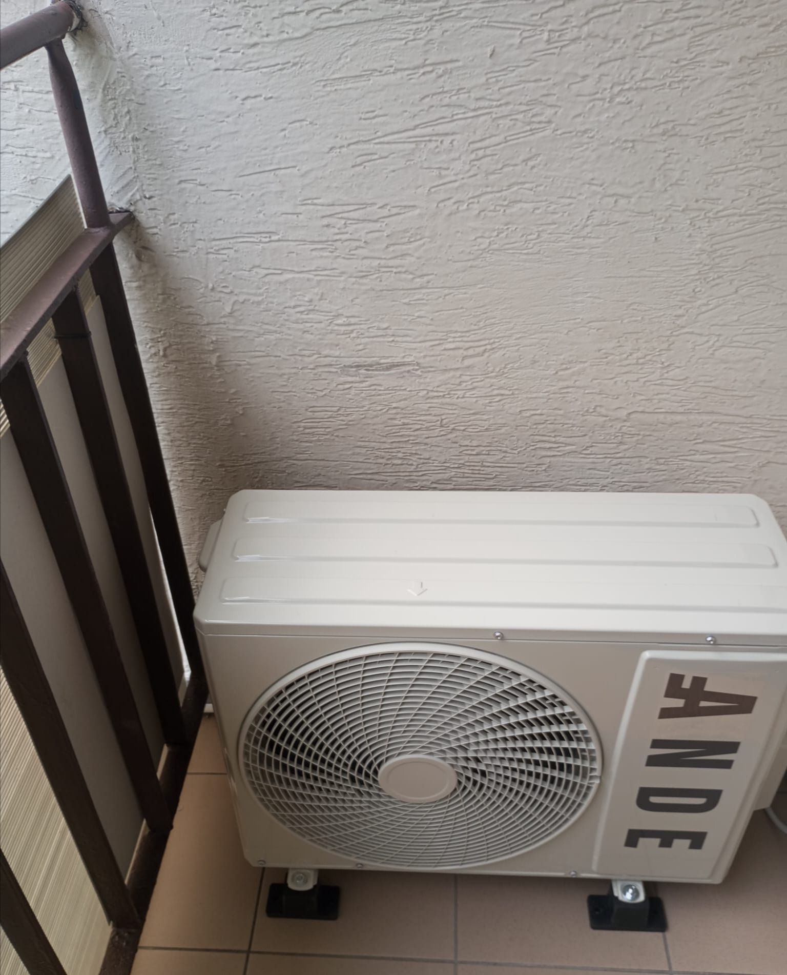 Zewnętrzna jednostka klimatyzacji marki ANDE zamontowana na balkonie, widoczna z góry, z fragmentem balustrady po lewej stronie i teksturą ściany w tle.
