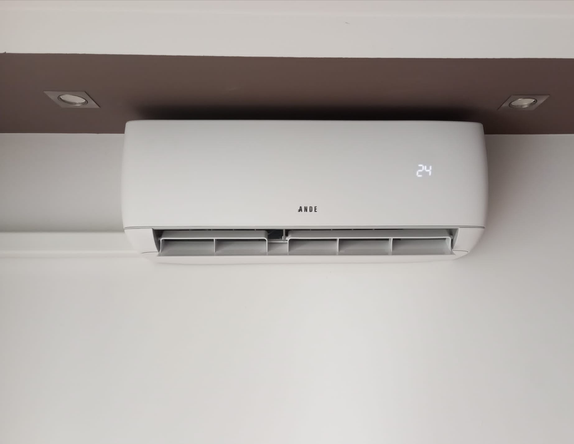Biała klimatyzacja marki ANDE zamontowana na ścianie, z wyświetlaczem wskazującym temperaturę 24 stopnie, oświetlona przez punktowe lampy sufitowe.