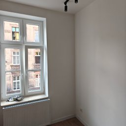 KAM-BUD Kamil Spieglanin - Pomalowane na biało puste pomieszczenie z drewnianą podłogą, białym oknem z widokiem na kamienicę i czarnym oświetleniem sufitowym.