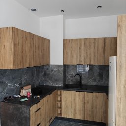 KAM-BUD Kamil Spieglanin - Nowoczesna kuchnia z drewnianymi szafkami, czarnym blatem i marmurowymi płytkami. Na blacie leżą okulary, torebka i puszka napoju.