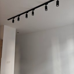 KAM-BUD Kamil Spieglanin - Czarny system szynowy z sześcioma reflektorami cylindrycznymi zamontowany na białym suficie w świeżo wyremontowanym pomieszczeniu z gładkimi, białymi ścianami i widocznym fragmentem podłogi...