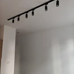 Czarny system szynowy z sześcioma reflektorami cylindrycznymi zamontowany na białym suficie w świeżo wyremontowanym pomieszczeniu z gładkimi, białymi ścianami i widocznym fragmentem podłogi...