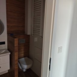 KAM-BUD Kamil Spieglanin - Widok z perspektywy wnętrza łazienki przez uchylone, białe drzwi, ukazujący fragment umywalki z szafką, podwieszaną toaletę z obudową imitującą drewno oraz grzejnik drabinkowy.