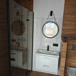 KAM-BUD Kamil Spieglanin - Nowoczesna łazienka z kabiną prysznicową typu walk-in z czarnymi profilami, okrągłym lustrem na sznurku, białą szafką pod umywalką i płytkami imitującymi drewno na ścianie.