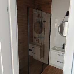 KAM-BUD Kamil Spieglanin - Nowoczesna łazienka z kabiną prysznicową typu walk-in z czarnymi profilami, białymi płytkami na ścianie i drewnopodobnymi płytkami na podłodze i części ściany, okrągłe lustro z podświetleniem LED...