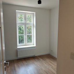 KAM-BUD Kamil Spieglanin - Pusty pokój po odświeżeniu, z drewnopodobną podłogą, białymi ścianami, grzejnikiem pod oknem i nowoczesną, czarną lampą sufitową.