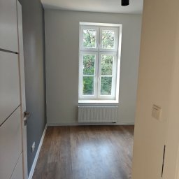 KAM-BUD Kamil Spieglanin - Pusty pokój po remoncie z drewnianą podłogą, grzejnikiem pod oknem i nowoczesnym oświetleniem sufitowym. Ściana pomalowana na szaro w kontrastowym zestawieniu z bielą okna i reszty ścian.