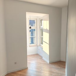 KAM-BUD Kamil Spieglanin - Pusty pokój po remoncie z drewnianą podłogą, białymi ścianami i oknem z widokiem na budynek, oświetlony naturalnym światłem.