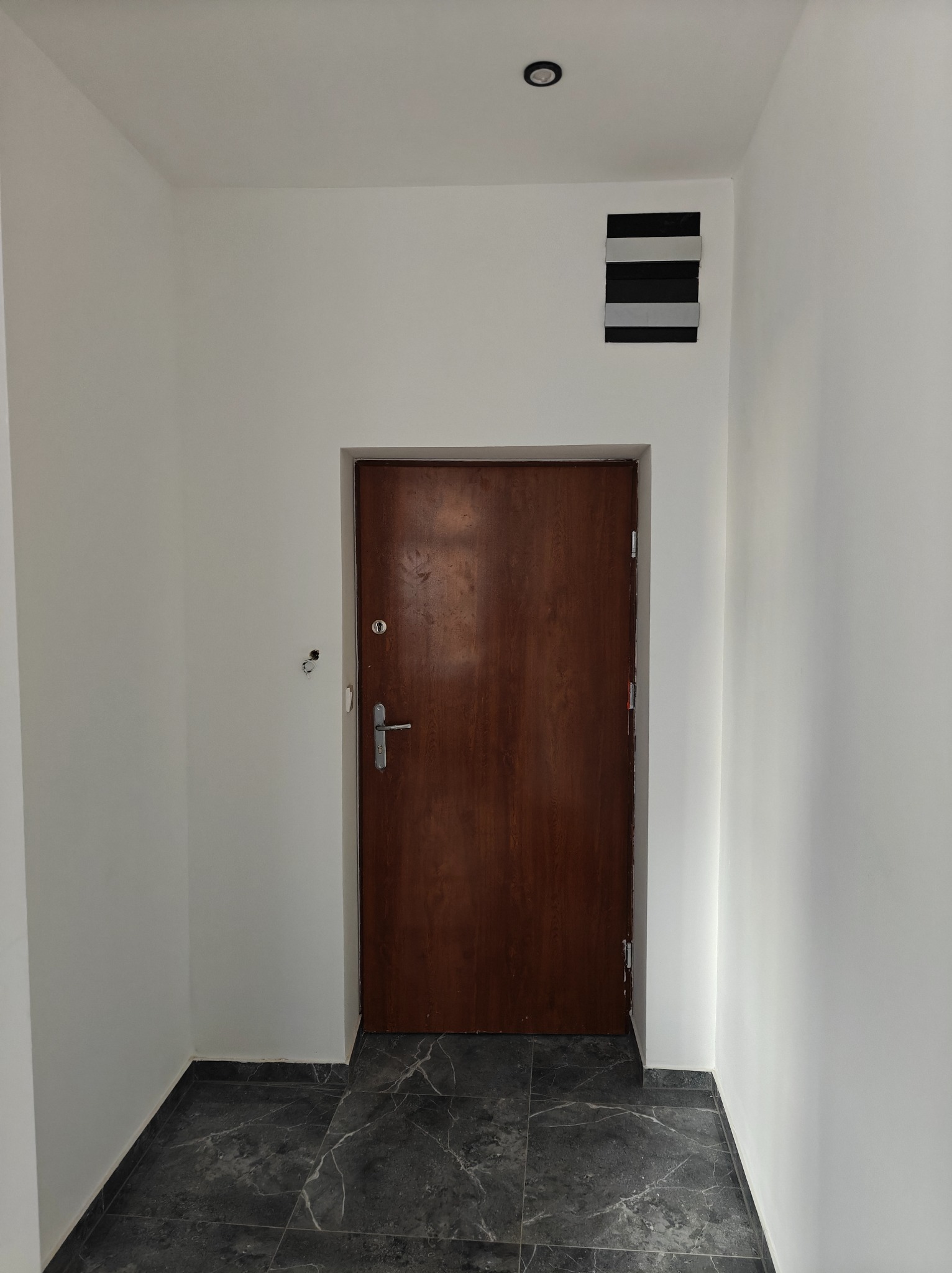Wnętrze korytarza po remoncie: białe ściany, ciemna marmurowa podłoga, drewniane drzwi wejściowe z metalową klamką, kratka wentylacyjna pod sufitem.