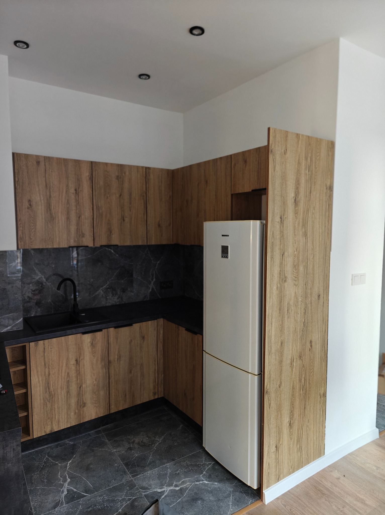 Nowoczesna kuchnia z drewnopodobnymi szafkami, czarnym blatem i marmurową ścianą nad zlewem, uzupełniona białą lodówką w zabudowie.