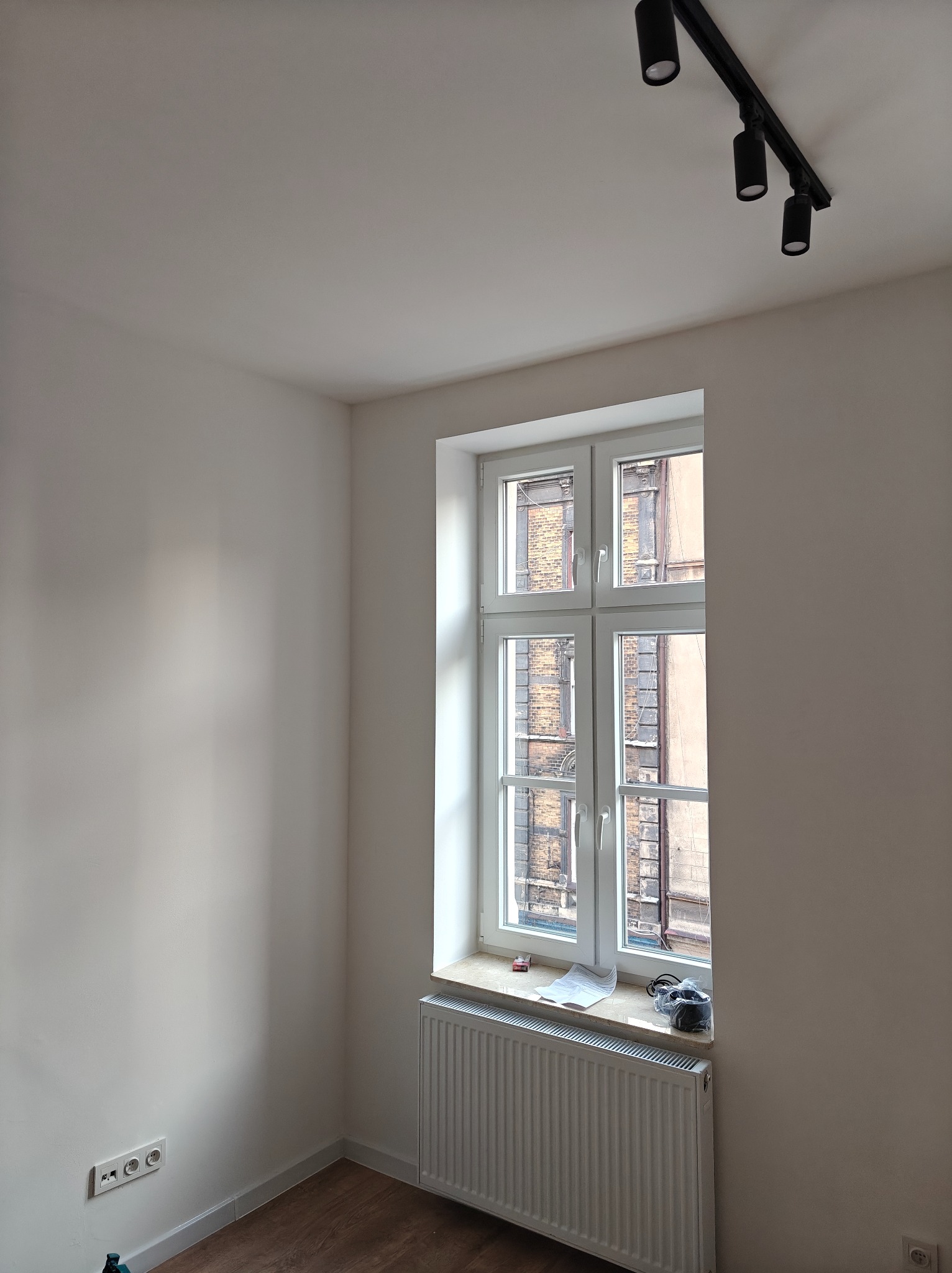 Wnętrze po odświeżeniu ścian na biało, widoczne gładkie ściany, okno z białymi ramami, grzejnik, listwa z trzema reflektorami na suficie i gniazdka elektryczne przy podłodze.