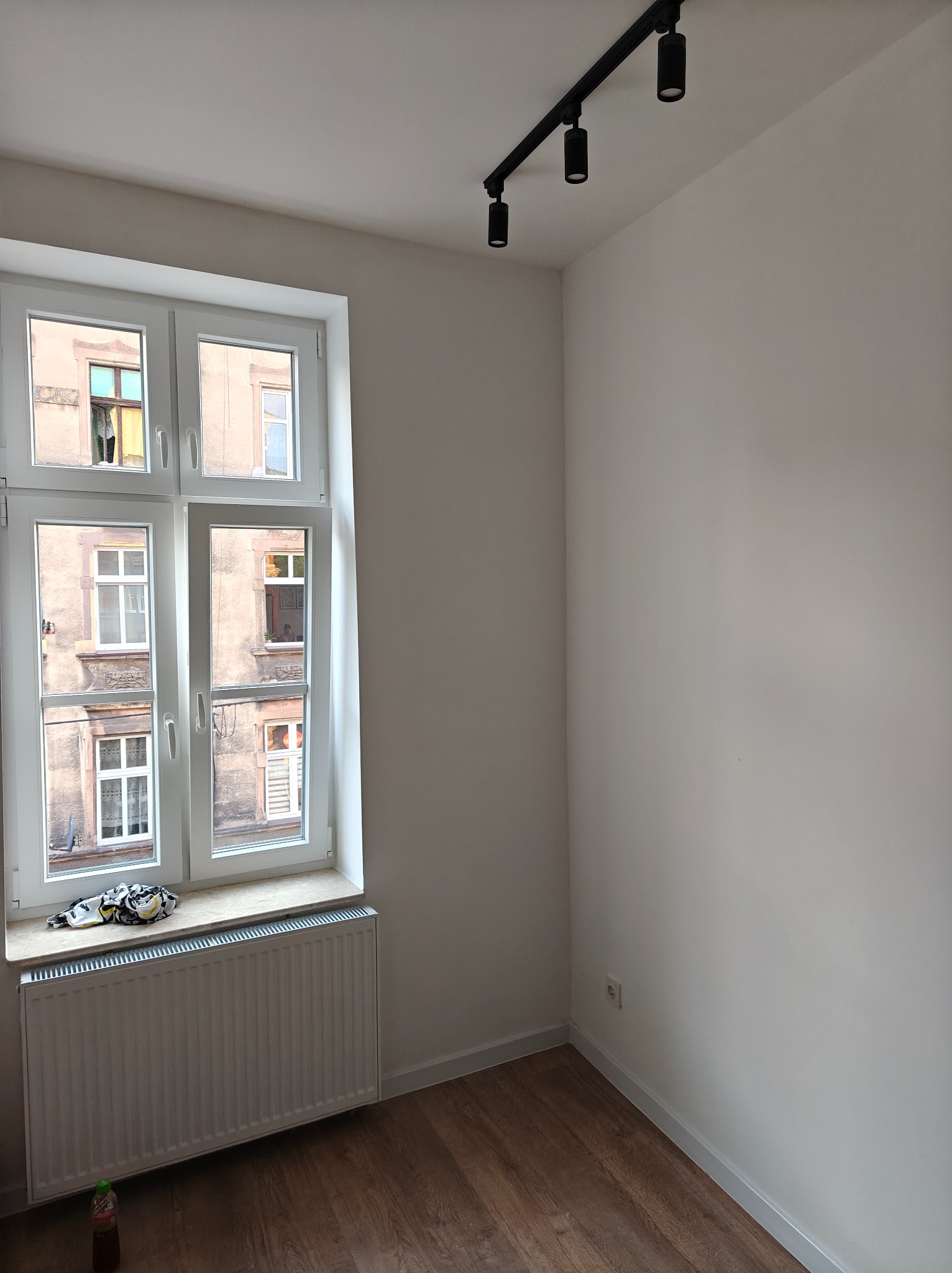 Pomalowane na biało puste pomieszczenie z drewnianą podłogą, białym oknem z widokiem na kamienicę i czarnym oświetleniem sufitowym.