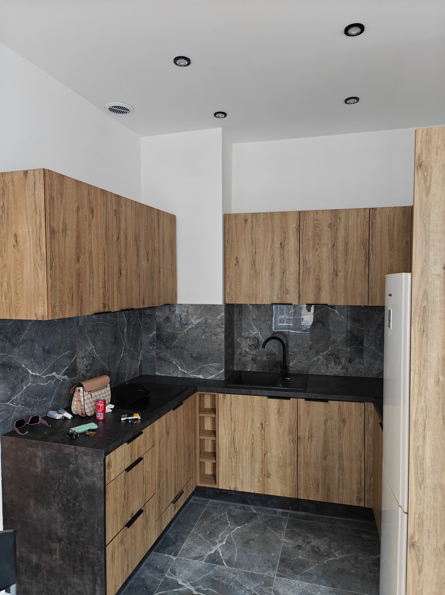 Nowoczesna kuchnia z drewnianymi szafkami, czarnym blatem i marmurowymi płytkami. Na blacie leżą okulary, torebka i puszka napoju.