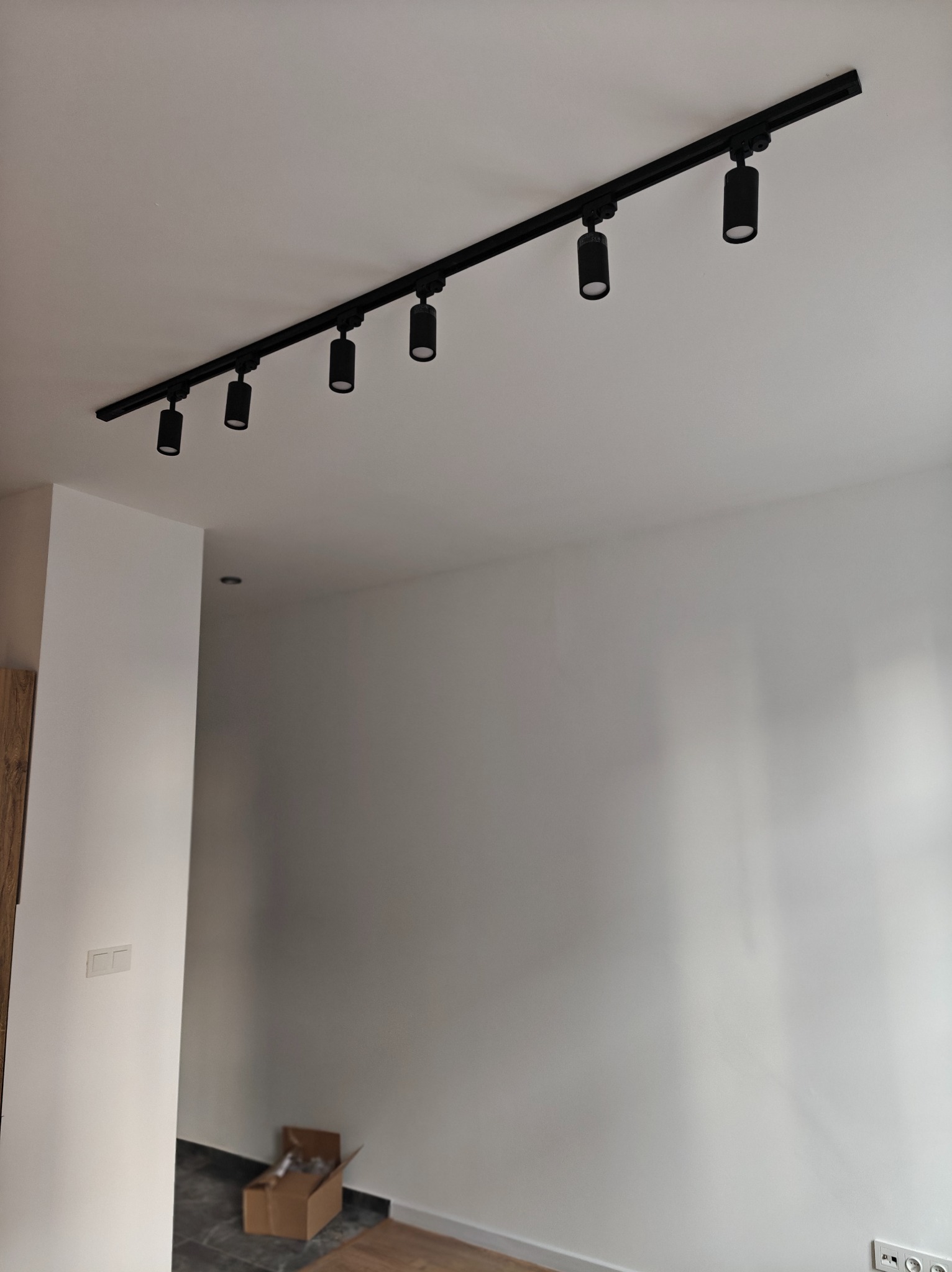 Czarny system szynowy z sześcioma reflektorami cylindrycznymi zamontowany na białym suficie w świeżo wyremontowanym pomieszczeniu z gładkimi, białymi ścianami i widocznym fragmentem podłogi...