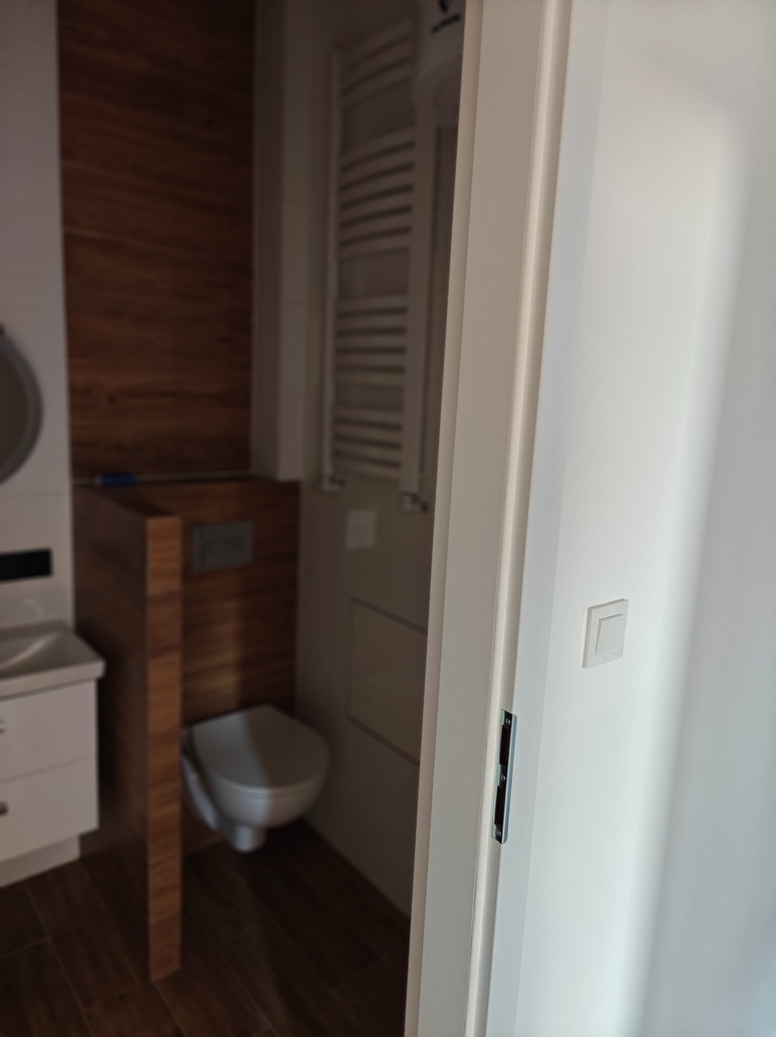 Widok z perspektywy wnętrza łazienki przez uchylone, białe drzwi, ukazujący fragment umywalki z szafką, podwieszaną toaletę z obudową imitującą drewno oraz grzejnik drabinkowy.