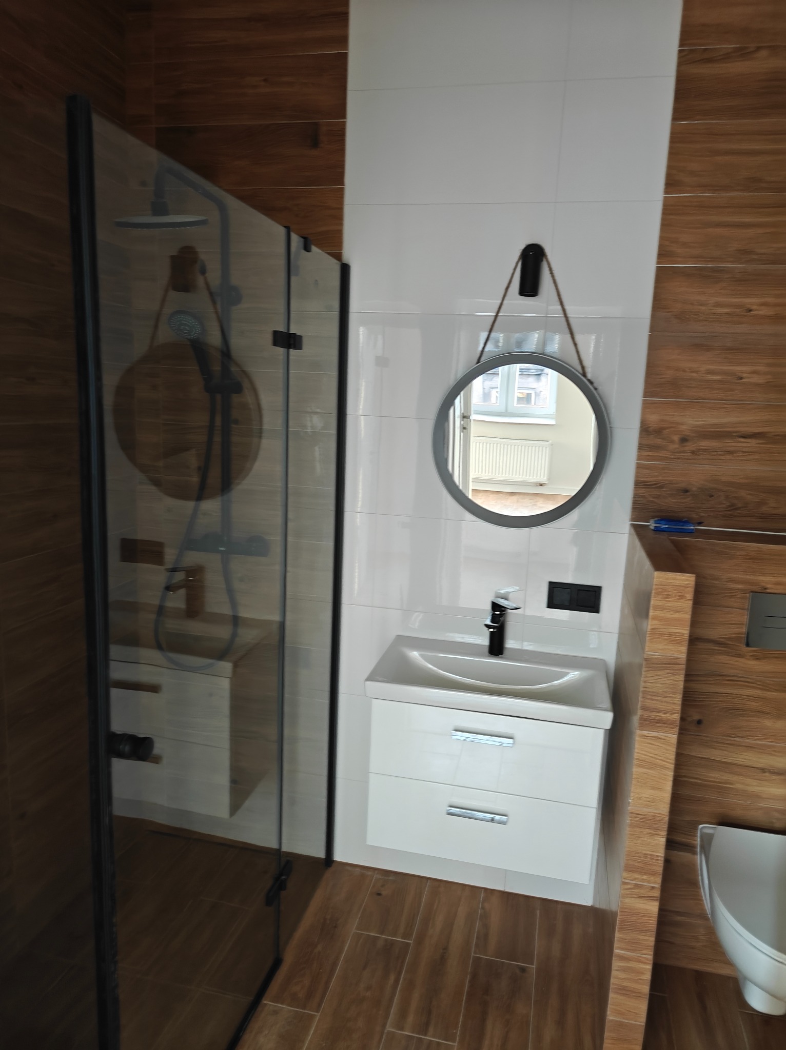 Nowoczesna łazienka z kabiną prysznicową typu walk-in z czarnymi profilami, okrągłym lustrem na sznurku, białą szafką pod umywalką i płytkami imitującymi drewno na ścianie.
