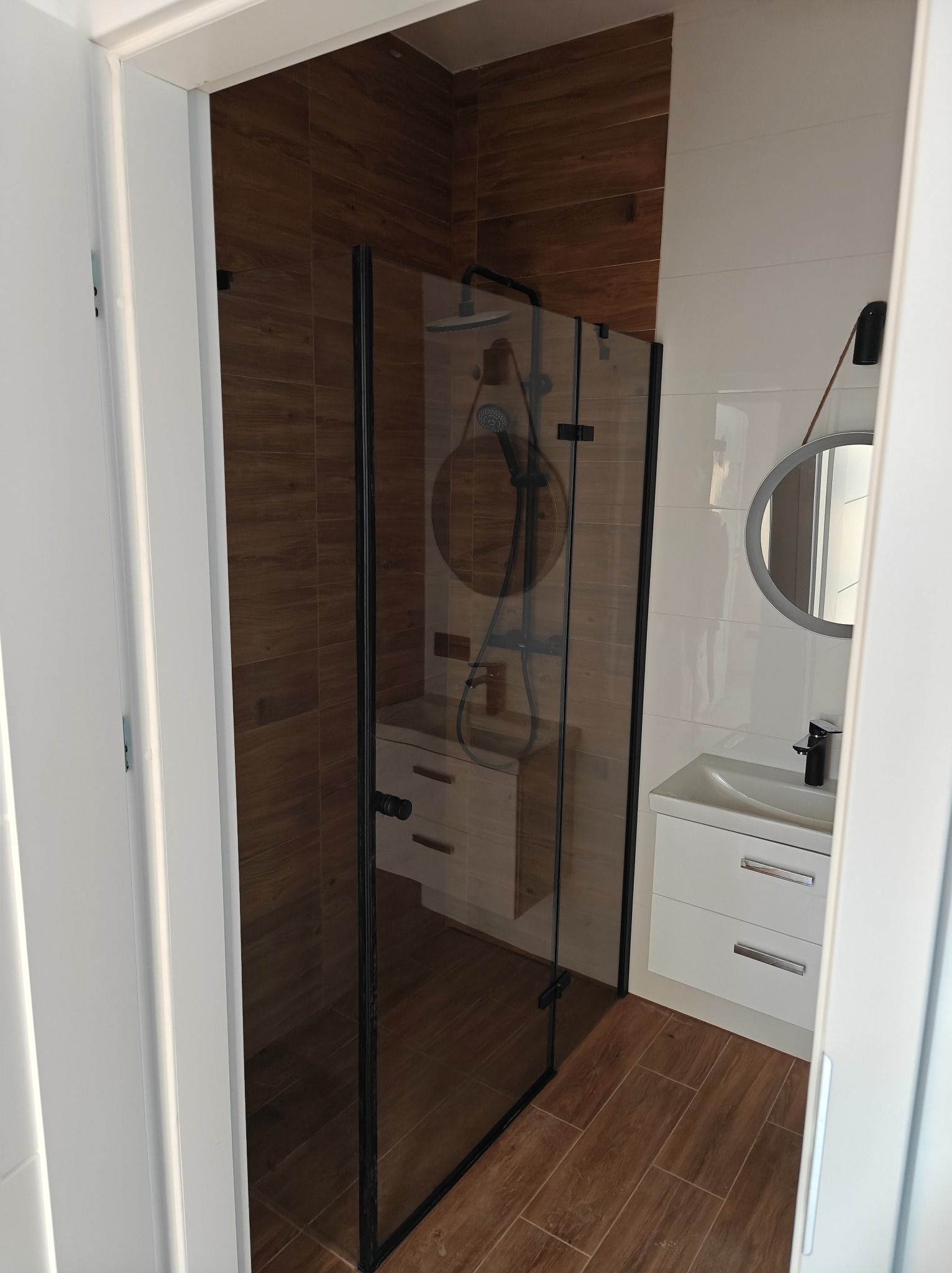 Nowoczesna łazienka z kabiną prysznicową typu walk-in z czarnymi profilami, białymi płytkami na ścianie i drewnopodobnymi płytkami na podłodze i części ściany, okrągłe lustro z podświetleniem LED...