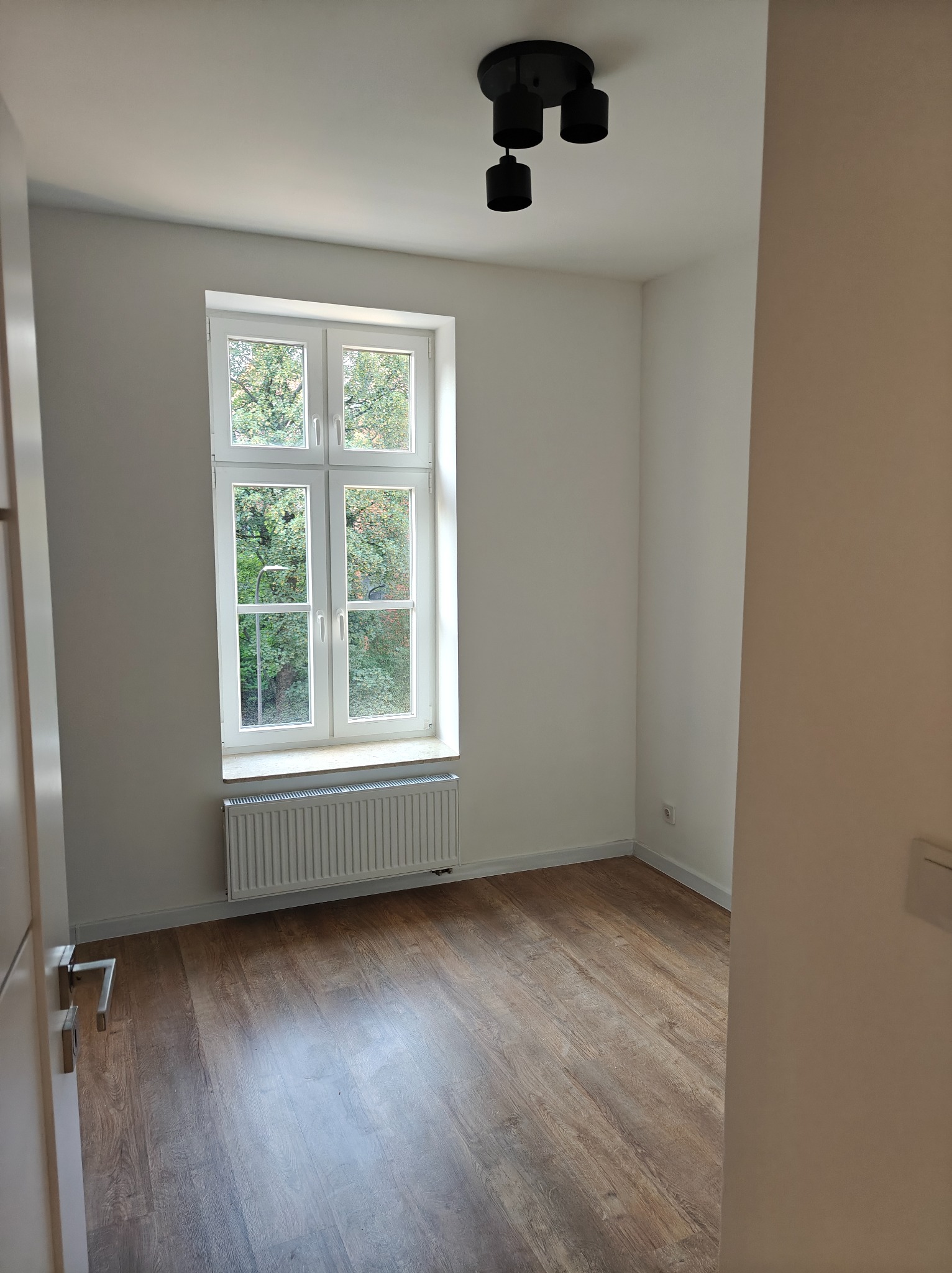 Pusty pokój po odświeżeniu, z drewnopodobną podłogą, białymi ścianami, grzejnikiem pod oknem i nowoczesną, czarną lampą sufitową.