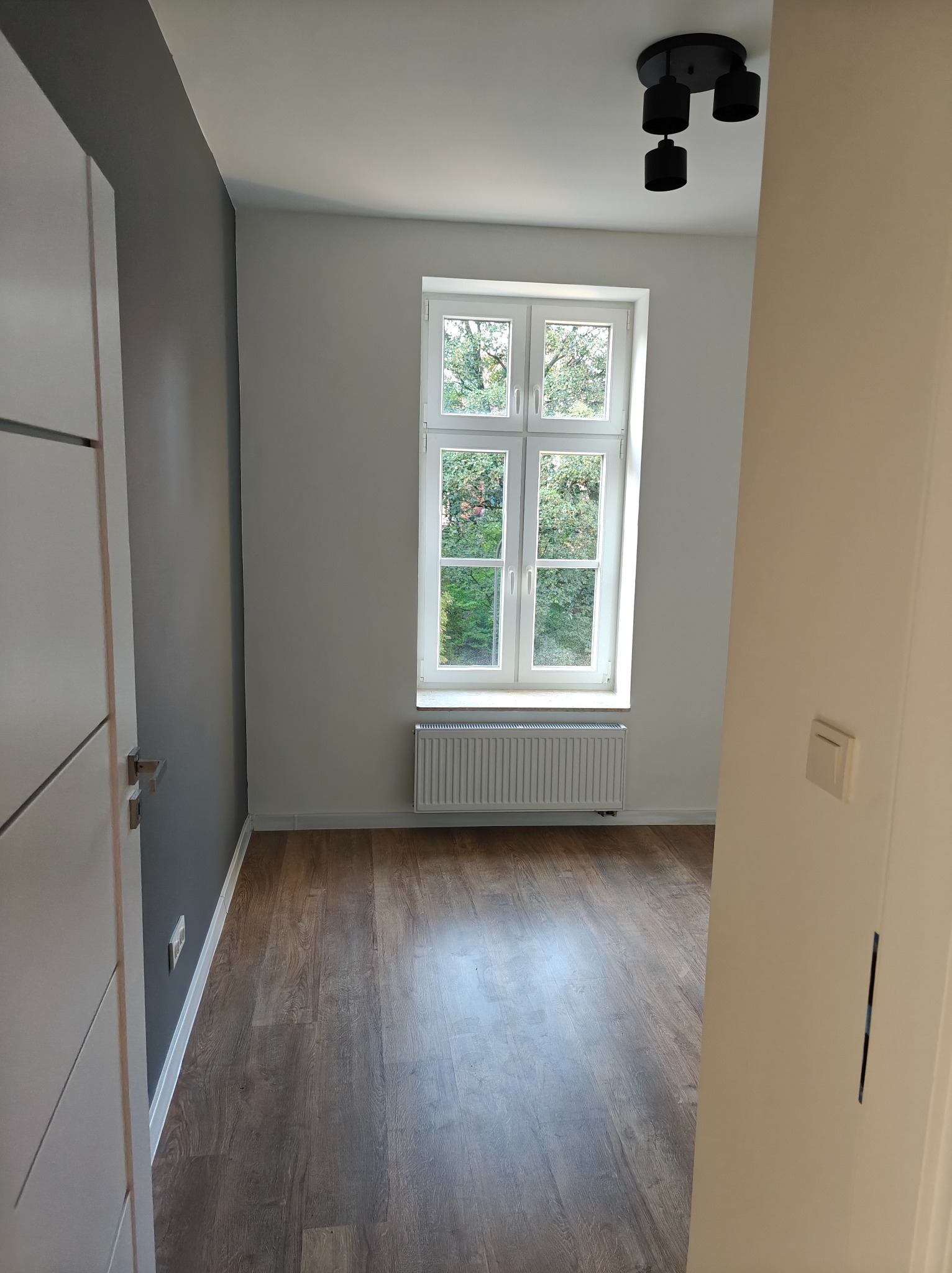 Pusty pokój po remoncie z drewnianą podłogą, grzejnikiem pod oknem i nowoczesnym oświetleniem sufitowym. Ściana pomalowana na szaro w kontrastowym zestawieniu z bielą okna i reszty ścian.