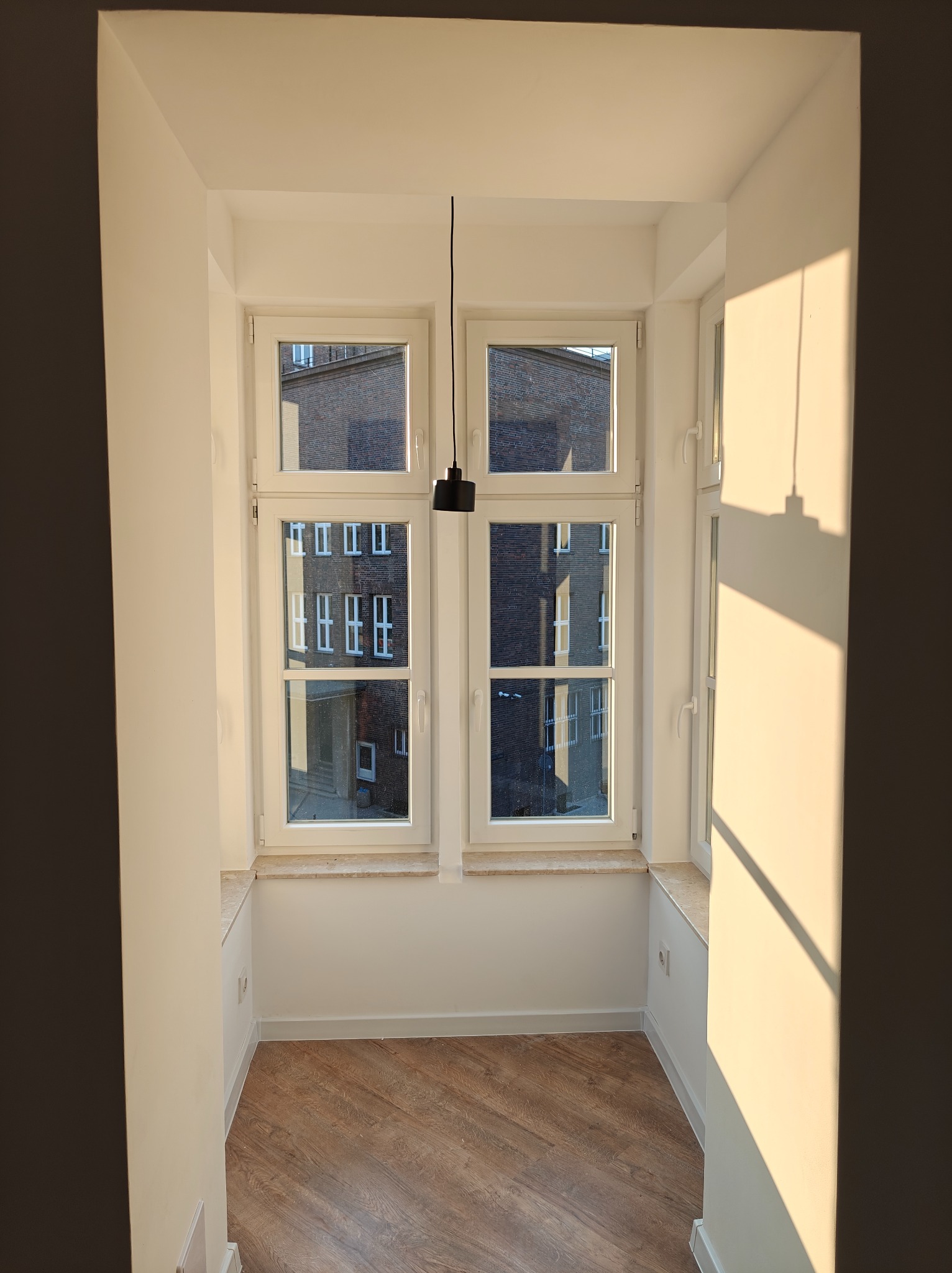 Wnęka okienna po remoncie z nową podłogą imitującą drewno, świeżo pomalowane ściany na biało, widoczne okno z budynkiem w tle i cień padający na ścianę.