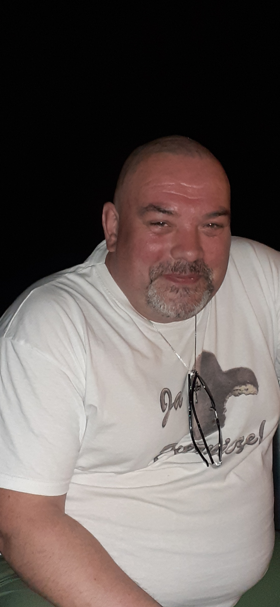 Portret mężczyzny z krótką brodą w białej koszulce z nadrukiem, na szyi wisi łańcuszek z okularami. Czarne tło. Uśmiechnięty, bezpośrednie spojrzenie.