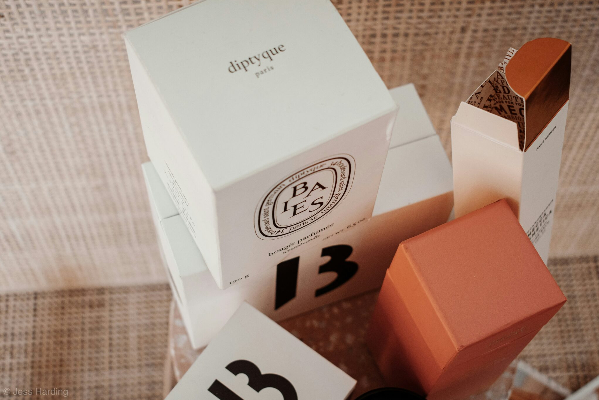 Zestaw eleganckich, markowych opakowań kosmetycznych w odcieniach bieli i terakoty, ułożonych na jutowym tle, z widocznym logotypem 'diptyque paris' i numerem '13'.