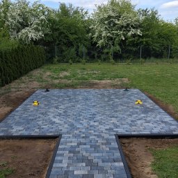 Marcin Karpiński - Ułożona kostka brukowa w kształcie litery T na trawniku, z czarnym obramowaniem i żółtymi narzędziami, gotowa do dalszych prac wykończeniowych.