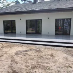 Nowoczesny taras z szarych płyt, z trzema stopniami i rampą, przylegający do białego budynku z trzema oknami. Na pierwszym planie piaszczyste podłoże.