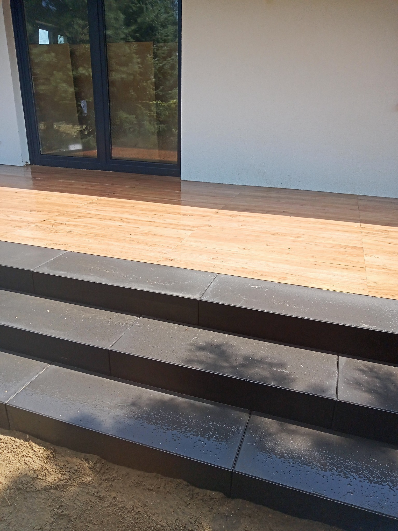 Nowoczesne czarne schody z płytek prowadzące do drewnopodobnego tarasu przy białej ścianie budynku z dużym oknem balkonowym.