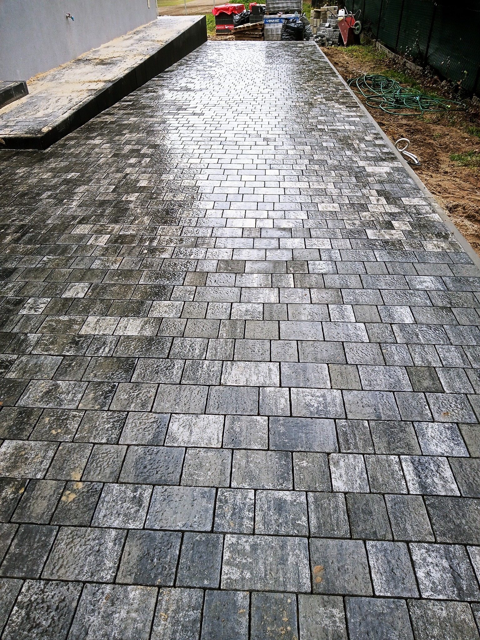 Ułożona, mokra kostka brukowa w odcieniach szarości prowadząca wzdłuż szarego budynku, widoczne narzędzia i materiały budowlane w tle.