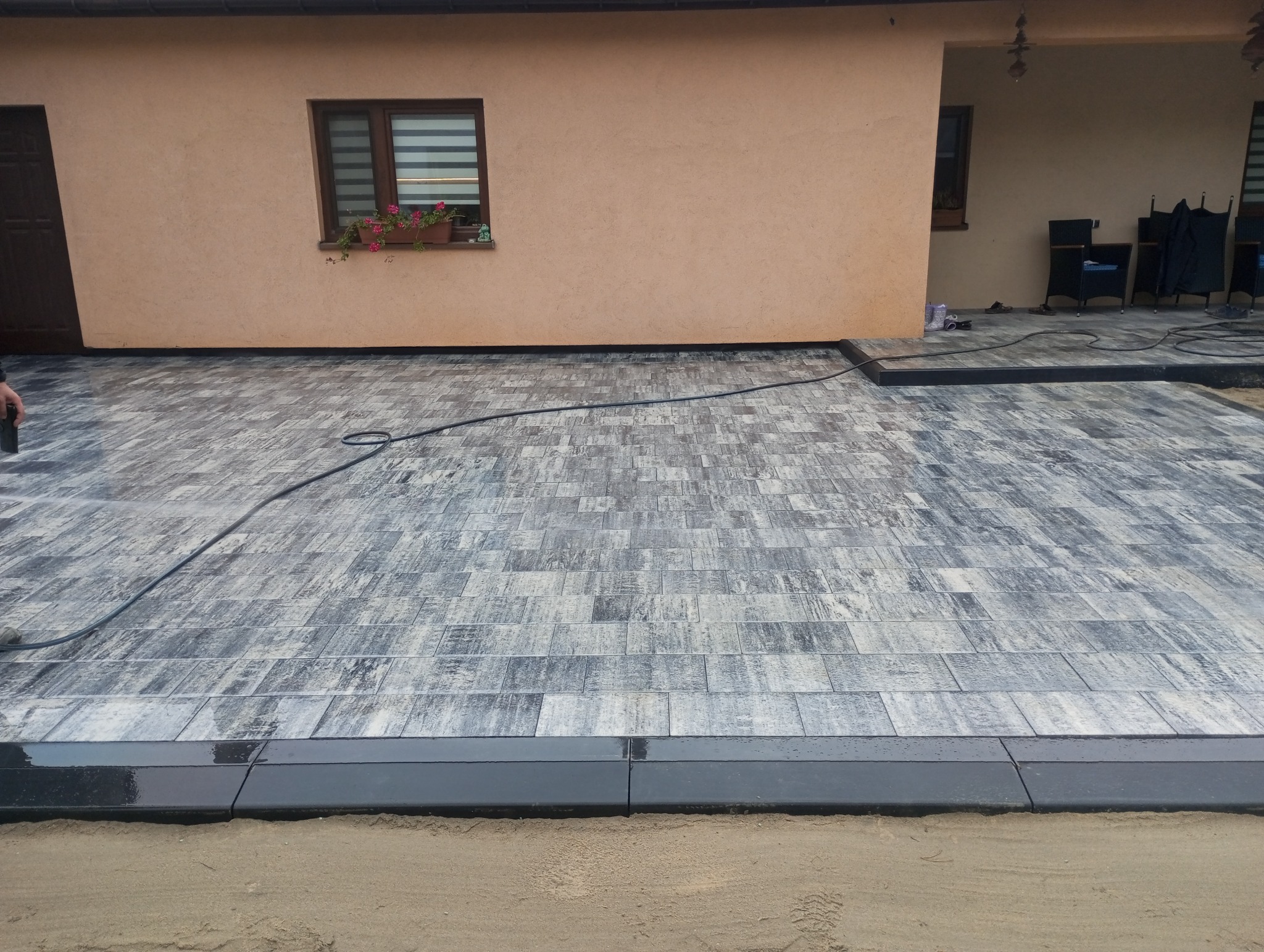 Świeżo ułożona kostka brukowa w odcieniach szarości z czarnym obrzeżem, mokra po myciu, widok na elewację domu z oknem i kwiatami.