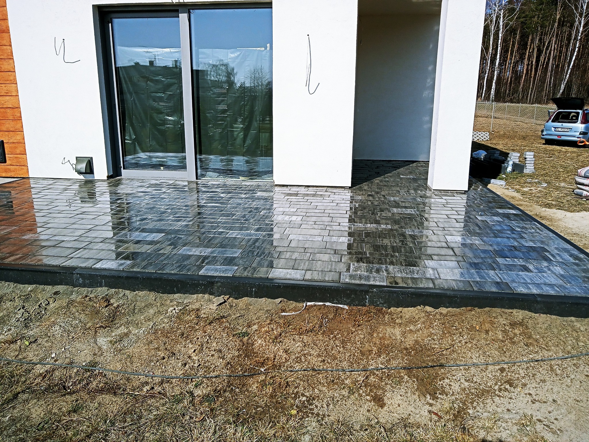 Świeżo ułożona, mokra kostka brukowa w odcieniach szarości przed nowoczesnym domem z białymi ścianami i dużym oknem, z widocznym fragmentem ogrodu w tle.
