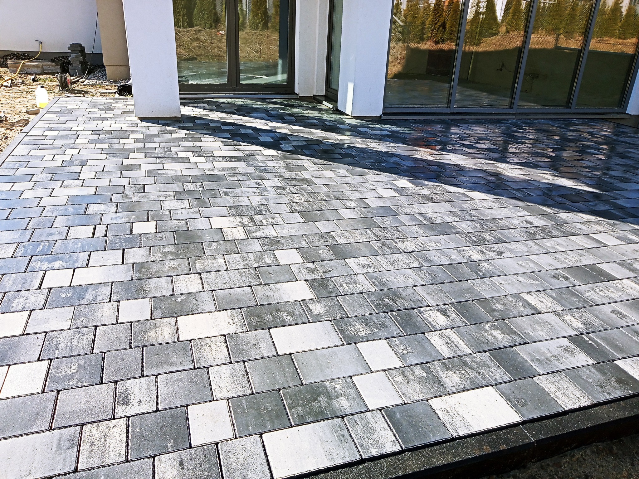 Ułożona kostka brukowa w odcieniach szarości i bieli, przylegająca do nowoczesnego budynku z dużymi oknami, widoczne lekkie zabrudzenia i piasek na obrzeżach.