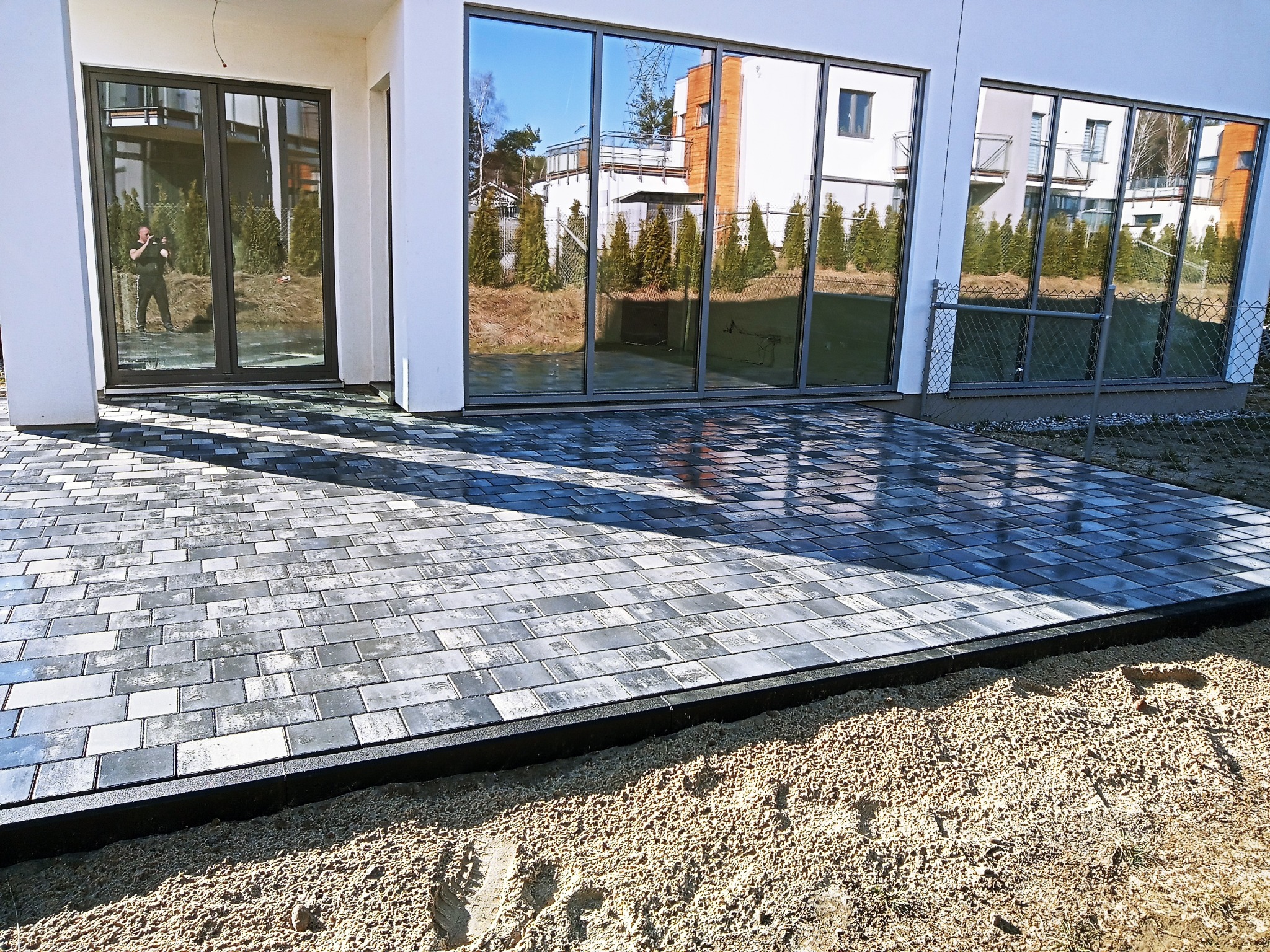 Ułożona szara kostka brukowa przed nowoczesnym domem z dużymi oknami, widoczne przygotowane podłoże z piasku i krawężnik.