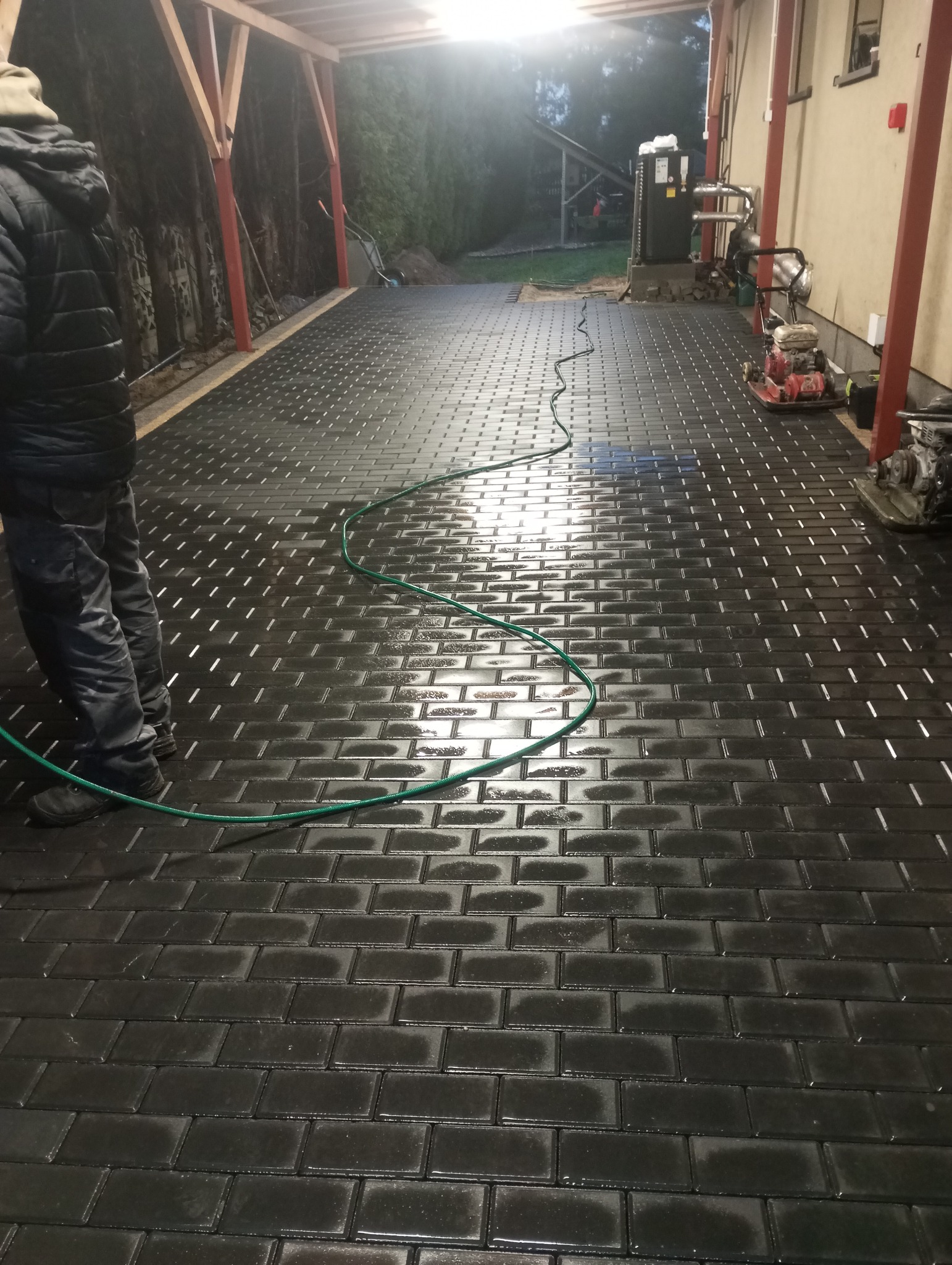 Świeżo ułożona, mokra kostka brukowa w kolorze antracytowym, ułożona pod zadaszeniem, z widocznym fragmentem osoby w kurtce i zielonym wężem ogrodowym, oraz sprzętem budowlanym w tle.