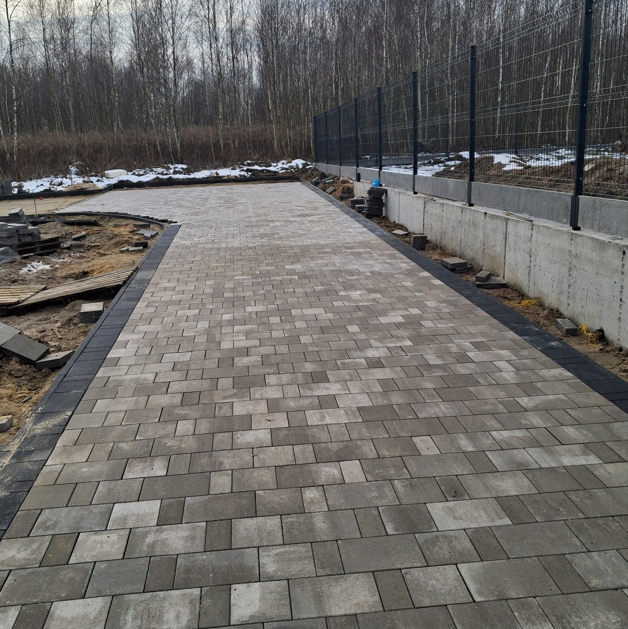 Ułożona kostka brukowa w odcieniach szarości, obrzeża z ciemnej kostki, widoczne fragmenty terenu przed budową ogrodzenia panelowego, drzewa w tle.
