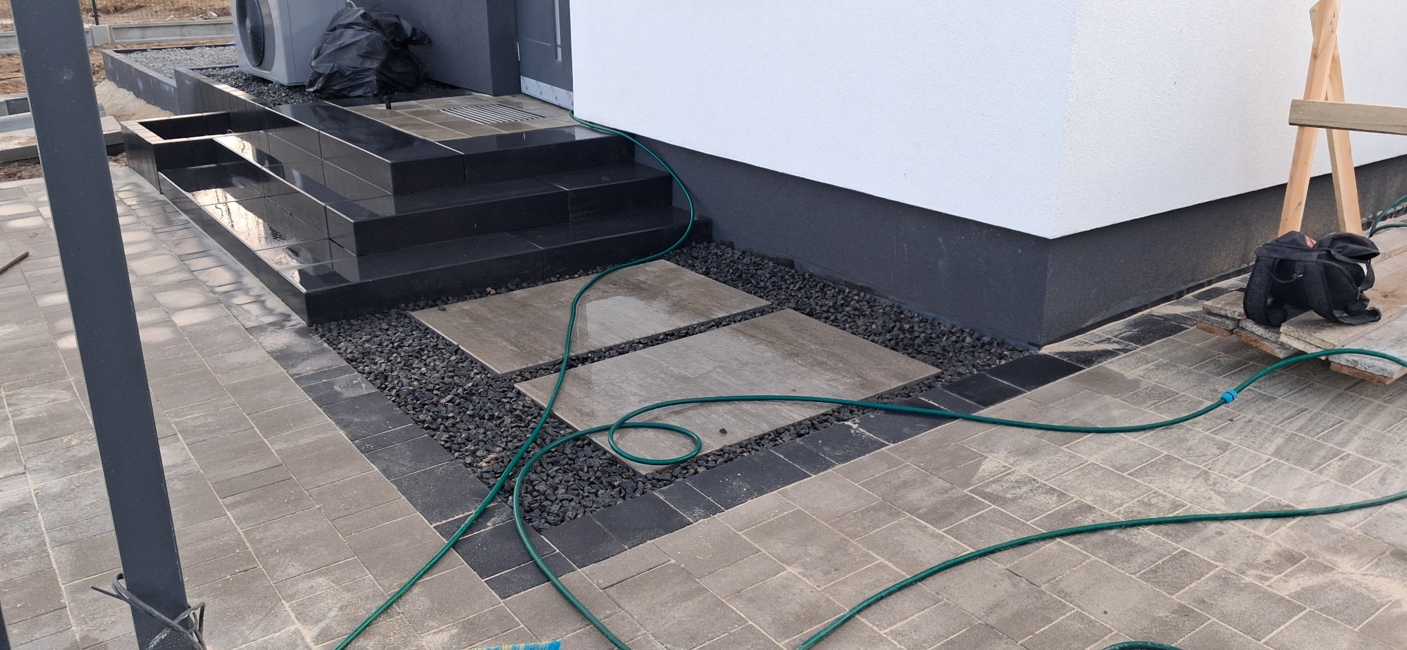 Ułożona kostka brukowa w odcieniach szarości, z czarnymi kamieniami ozdobnymi i schodami z ciemnego kamienia prowadzącymi do budynku; widoczny wąż ogrodowy i torba narzędziowa.