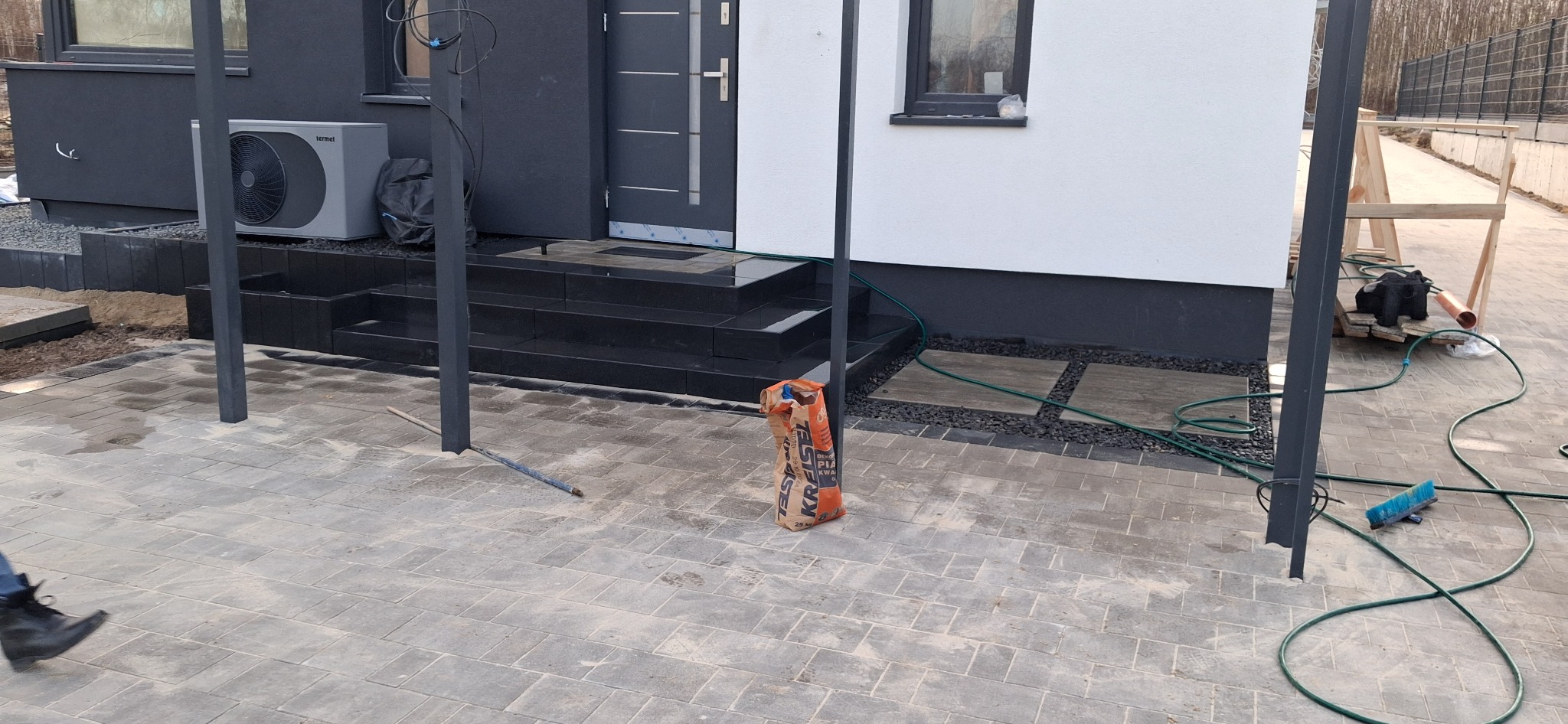 Świeżo ułożona, szara kostka brukowa prowadząca do nowoczesnego domu z czarnymi schodami i pompą ciepła, z workiem zaprawy KREISEL i narzędziami budowlanymi w tle.