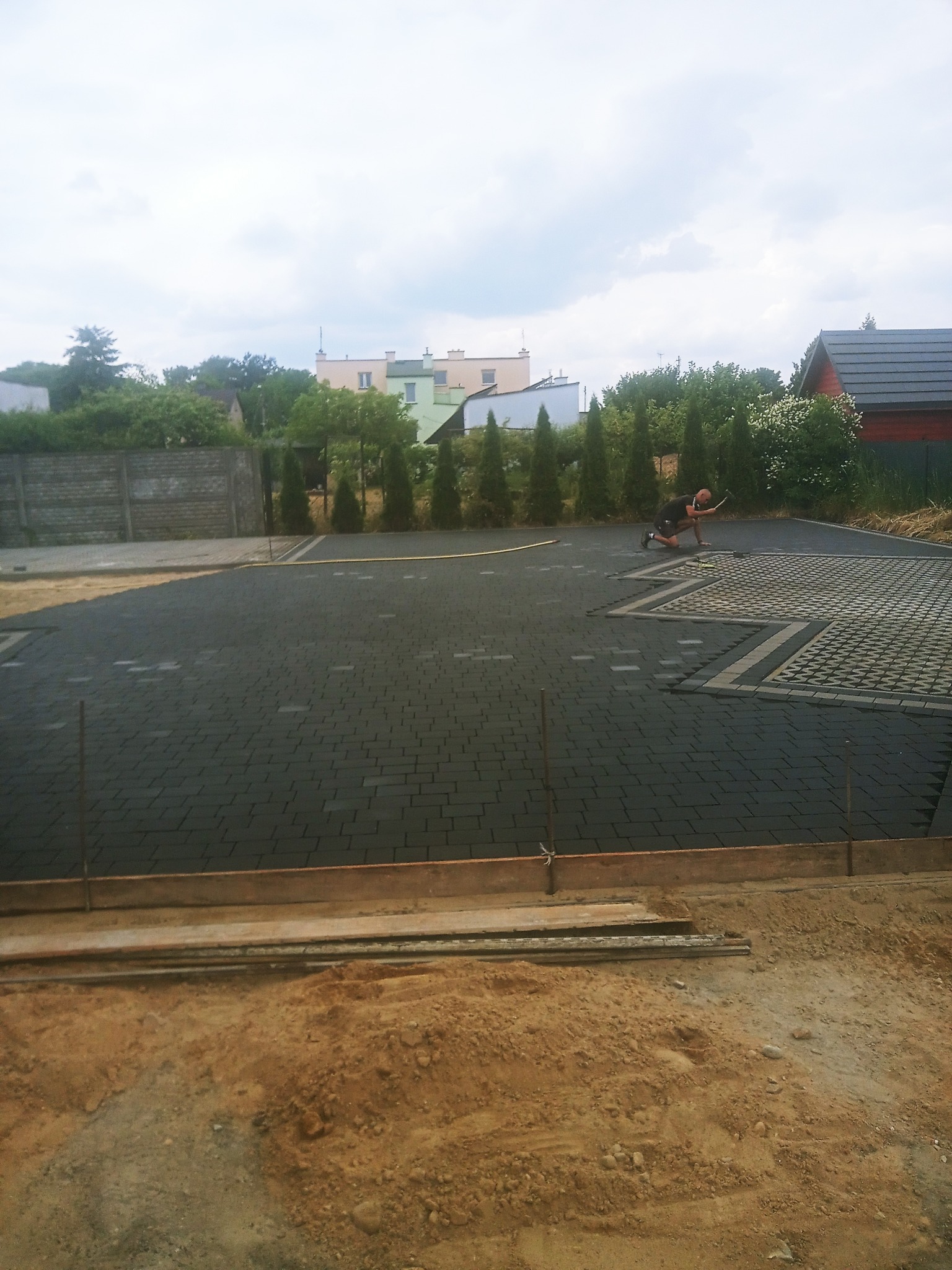 Układanie czarnej kostki brukowej na posesji, widoczny fragment ułożonej powierzchni z dekoracyjnym wzorem, pracownik z narzędziem w trakcie pracy, obrzeża w przygotowaniu.