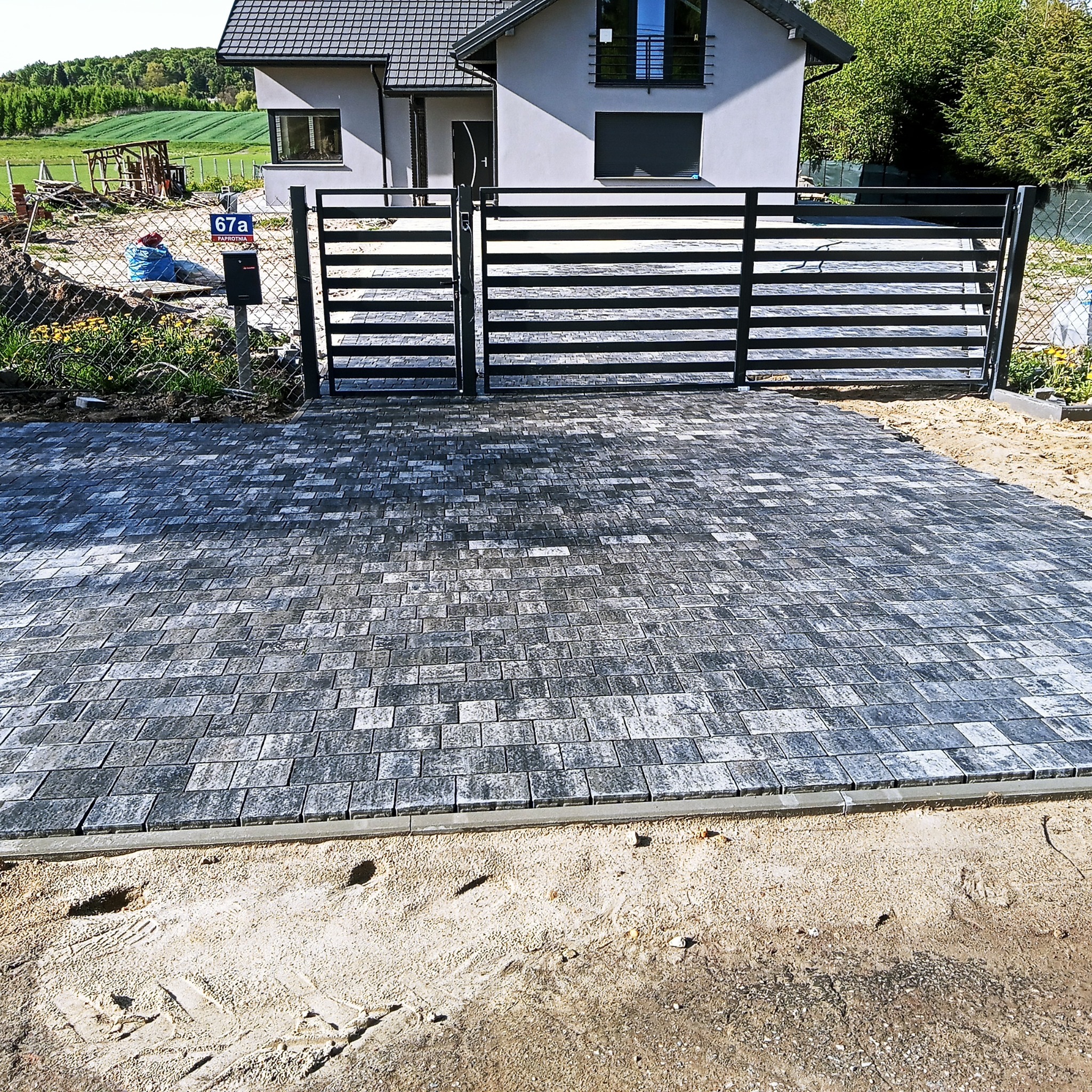 Nowoczesny, poziomy płot panelowy w kolorze antracytowym z bramą wjazdową, na tle szarego domu z ciemną dachówką i ułożonej kostki brukowej w odcieniach szarości.