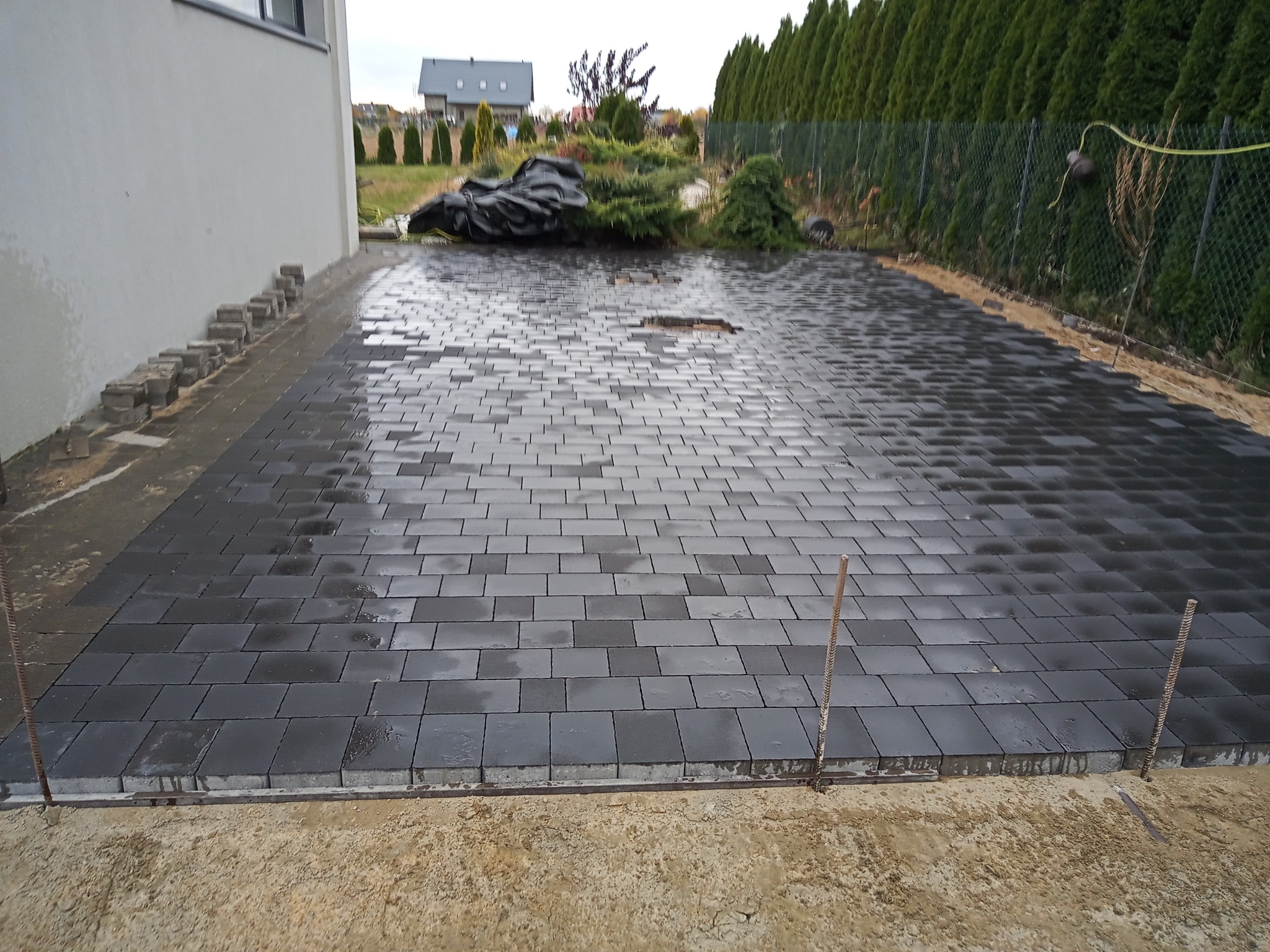 Układanie szarej kostki brukowej na posesji, widoczne stalowe pręty stabilizujące obrzeże, w tle dom i zieleń.