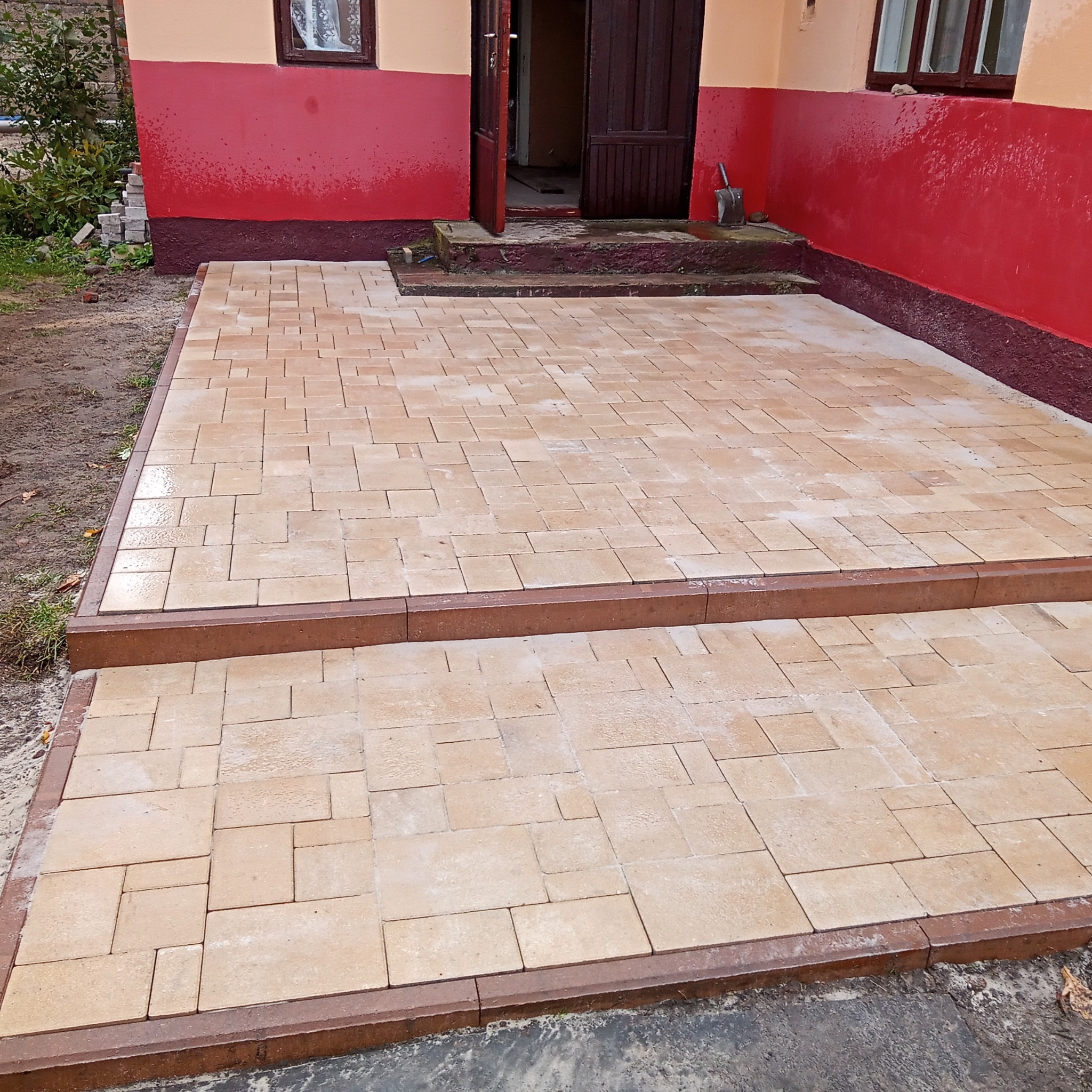 Nowo ułożona, jasna kostka brukowa prowadząca do wejścia domu z czerwono-beżową elewacją, widoczne dwa stopnie z obrzeżami w kolorze ciemnego brązu.