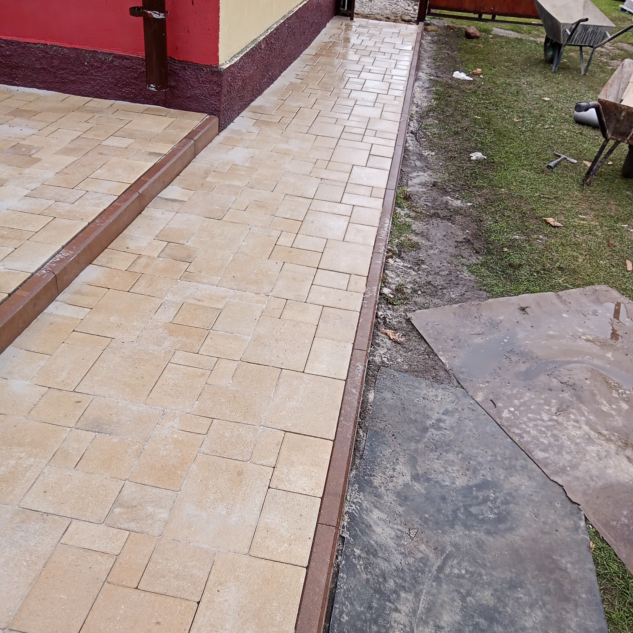 Świeżo ułożona, beżowa kostka brukowa tworząca ścieżkę z brązowym obramowaniem, prowadząca od budynku w stronę trawnika z widocznymi taczkami i narzędziami.