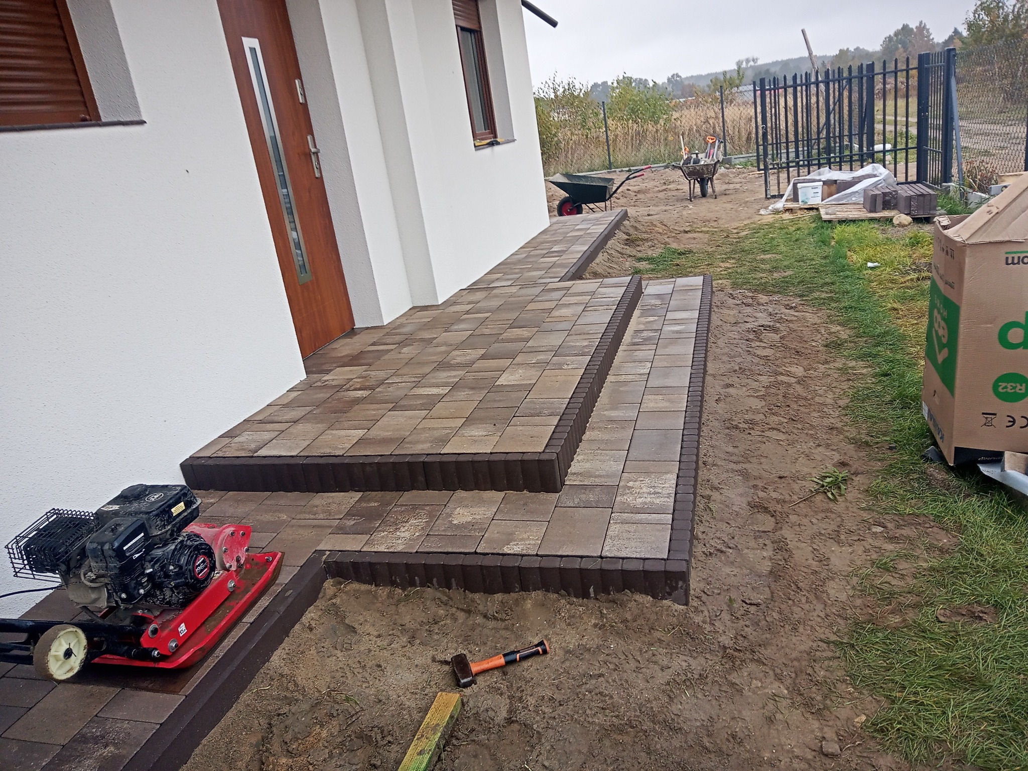 Ułożona kostka brukowa tworząca schody wejściowe do domu, z widoczną zagęszczarką w pobliżu i narzędziami na ziemi, w tle częściowo widoczne ogrodzenie posesji.