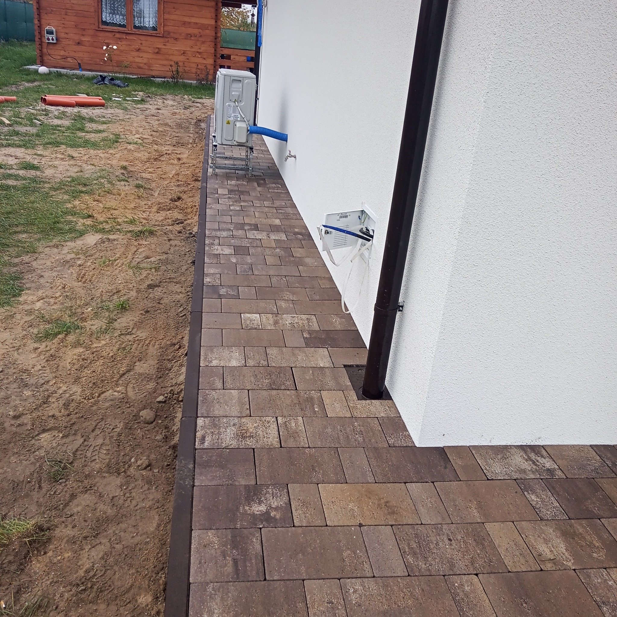 Ułożona kostka brukowa wzdłuż ściany budynku z widoczną rurą spustową i instalacją klimatyzacji na zewnątrz.