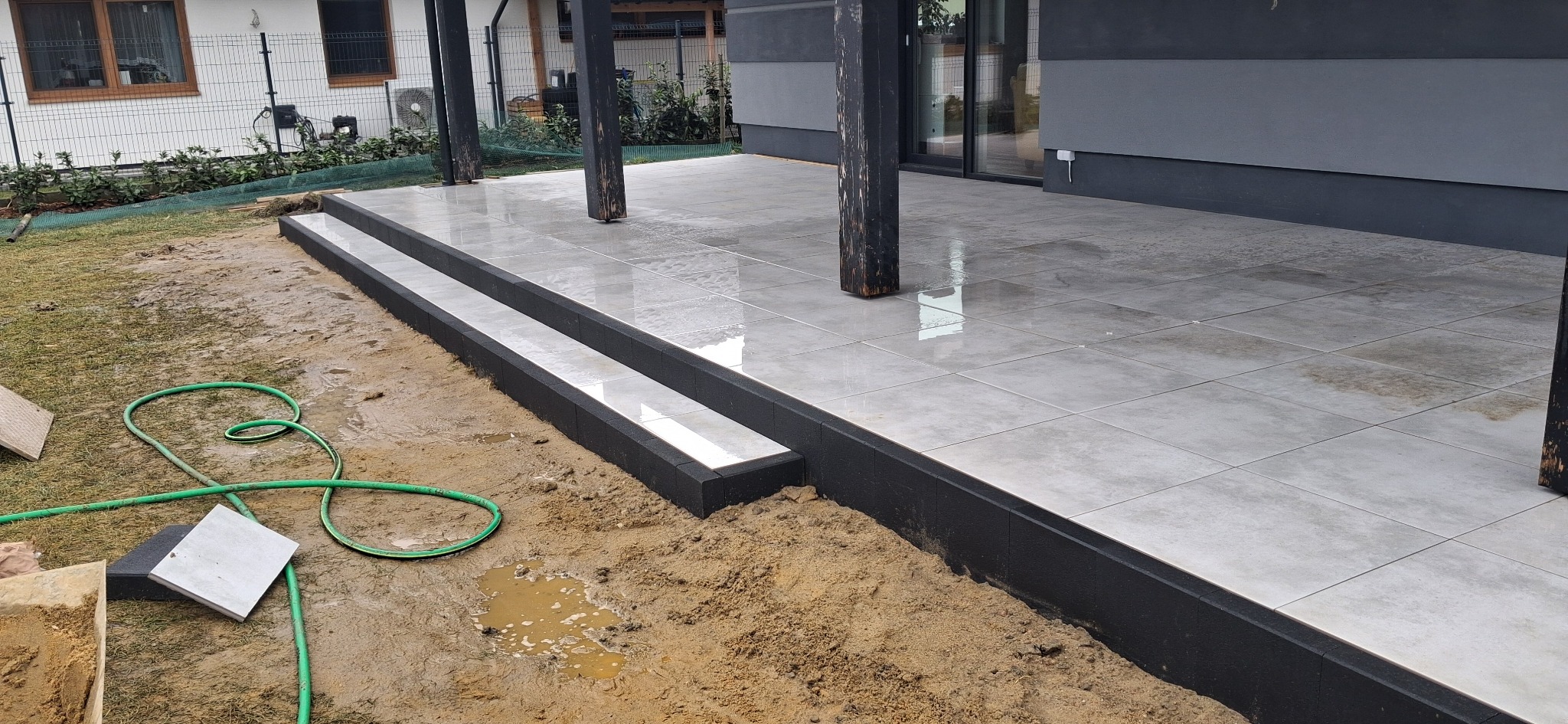 Ułożona, jasnoszara, kwadratowa kostka brukowa na tarasie z dwoma stopniami, obramowana czarnym obrzeżem. W tle widoczne drewniane słupy podtrzymujące zadaszenie i fragment domu z oknami.