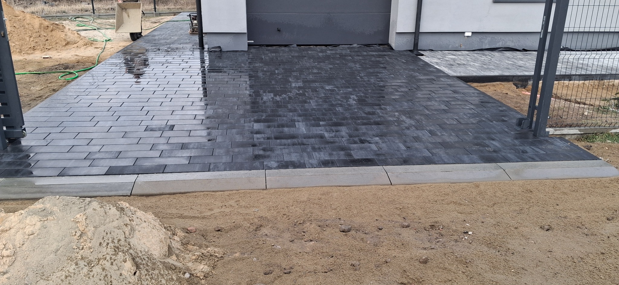 Świeżo ułożona, mokra, grafitowa kostka brukowa na podjeździe, obrzeża betonowe, widoczna brama wjazdowa i ogrodzenie panelowe w tle, obok piasek.